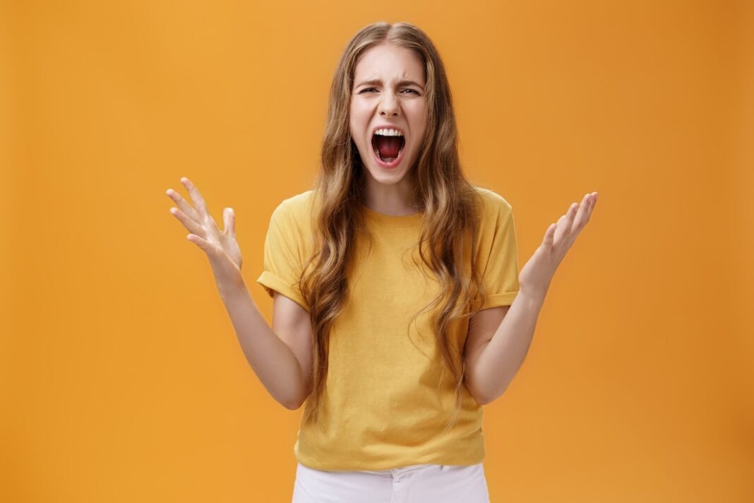 Studio shot of young woman during argument losing temper standing pressured and pissed yelling out loud from hate and anger gesturing with raised palms making furious face posing against orange wall